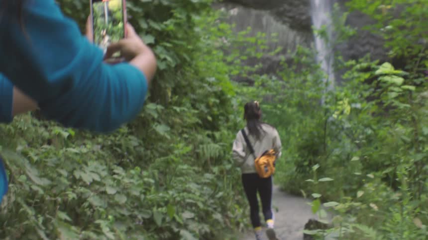 Woman taking a picture of another woman on a trail with a waterfall in the background