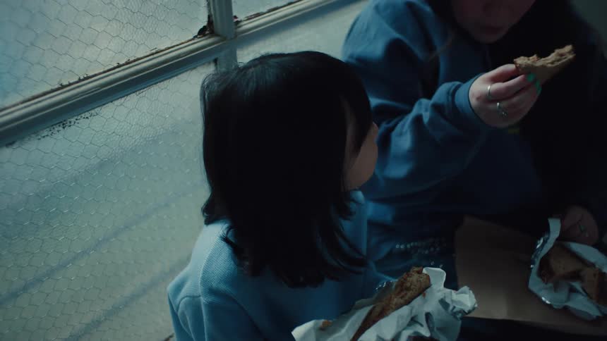 The video shows two people sitting indoors eating what appears to be sandwiches one person has long dark hair and is wearing a light blue sweatshirt the other person is wearing a blue hooded sweatshirt with a graphic design on the front and several rin