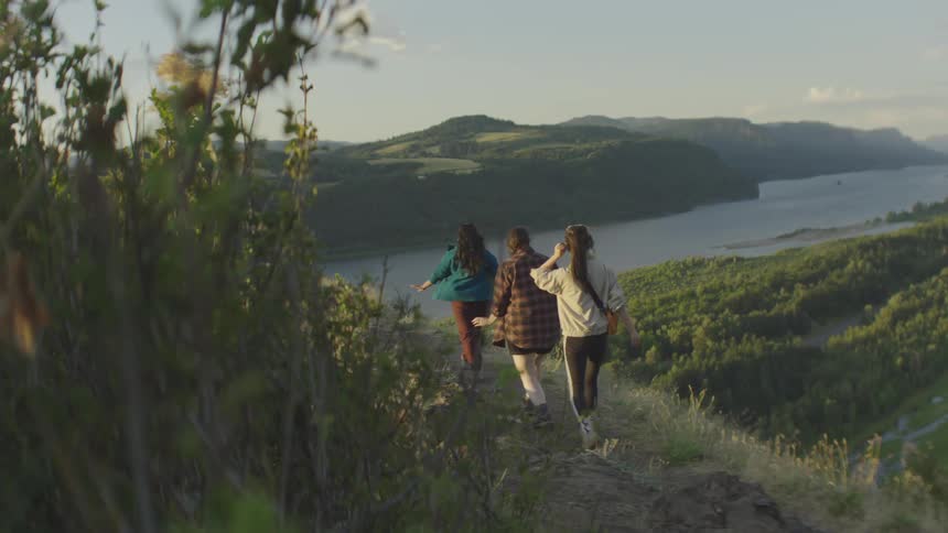 Three friends walking on a grassy hilltop overlooking a wide river and green hills with one of them raising her arms in the air