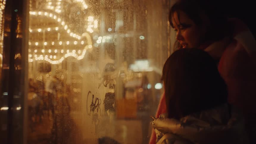 Woman and a child draw on a foggy glass window and in the background a lit carousel