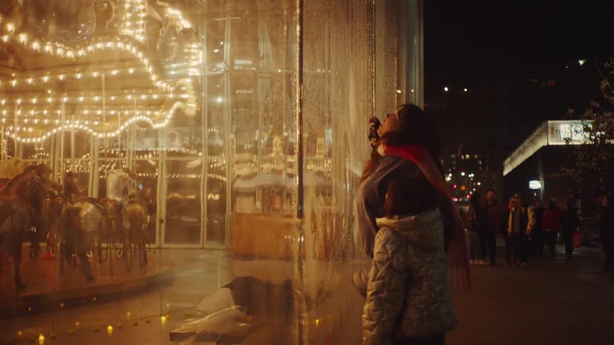 A mother and daughter stand in front of a wet glass window looking at a brightly lit merry go round and playing with condensation on the glass