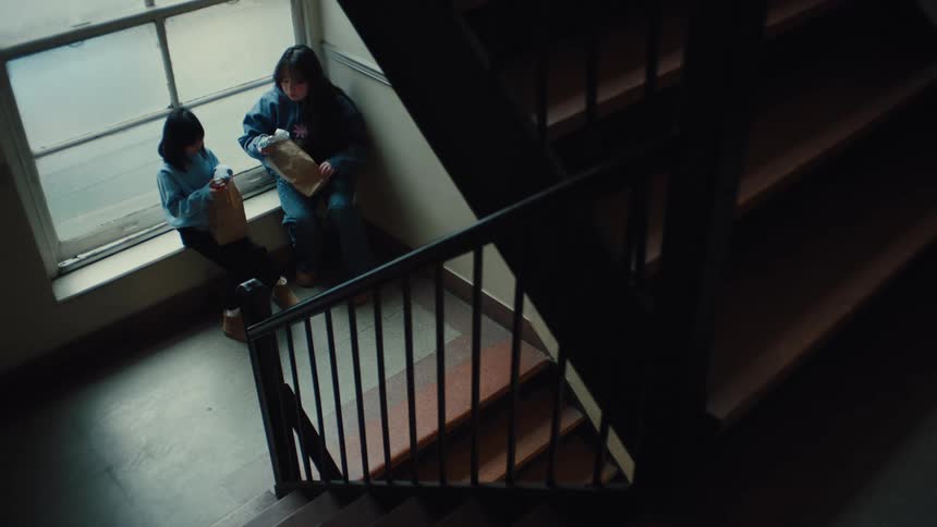 Two children quietly eat lunch together on a stairwell landing beside a sunlit window