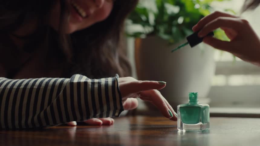 The video shows two people seated at a wooden table one person is applying green nail polish to the other persons hand which has a ring on a finger a potted plant is visible in the background