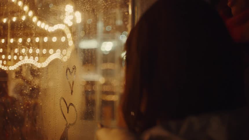 The video shows two people at night looking out a window with a blurredout reflection of a string of lights one person uses their finger to draw a heart and a letter on the foggy glass