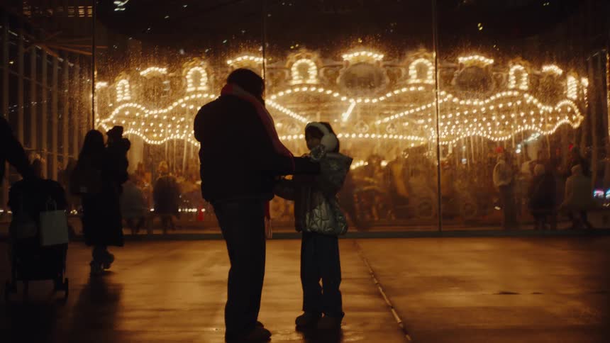 A parent and child are standing in front of a brightly lit carousel at night the parent is helping the child with their scarf while other people are visible in the background