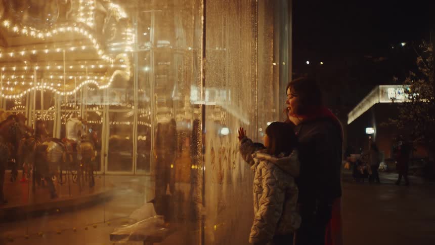A short video clip shows a woman and a child standing outside a carousel at night looking in the carousel is lit up with many light bulbs the woman has long dark hair and is wearing a dark jacket with a red scarf and the child is wearing a puffy jacke