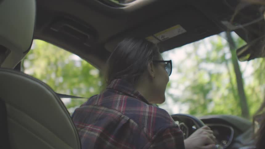 A woman wearing a red and blue plaid shirt and sunglasses drives a car through a forest briefly turning to smile at a passenger in the back seat