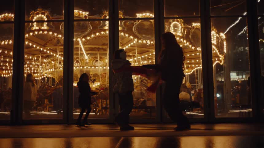 A mother and daughter play with a red scarf while standing in a dark room in front of a large window that shows a brightly lit moving merry go round