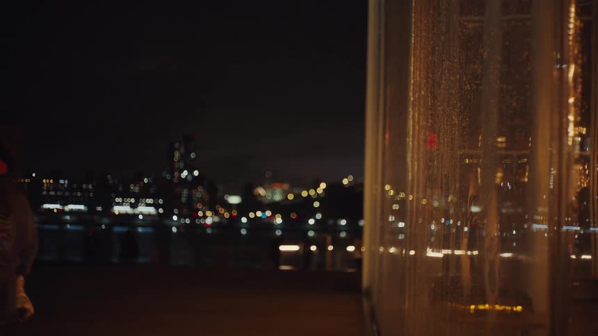 A young girl in a puffy gold jacket looks out a window at a dark cityscape with her hands pressed against the glass another person joins her and they smile as they look at the city lights together