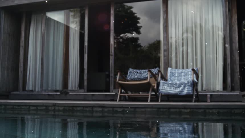 Water splashes near poolside lounge chairs in front of a modern house