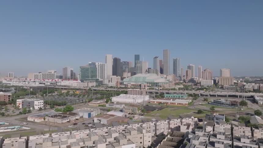 A short aerial video clip shows a cityscape under a clear blue sky in the foreground are many smaller buildings including a residential area with similarlooking rooftops the background is filled with a large cluster of modern skyscrapers the logo fi