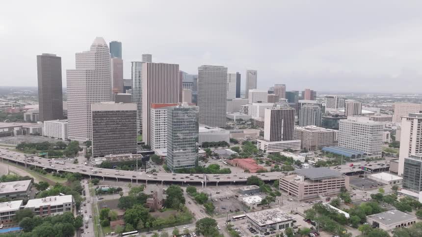 A large city skyline with numerous tall buildings and a busy highway is visible under a cloudy gray sky