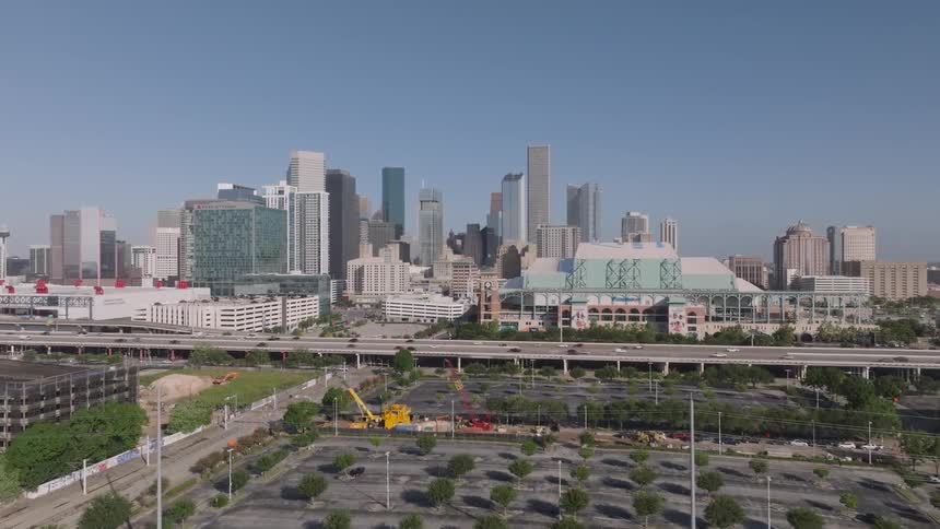 A large parking lot with small trees and a busy overpass is in the foreground of a city skyline with many tall skyscrapers under a clear blue sky