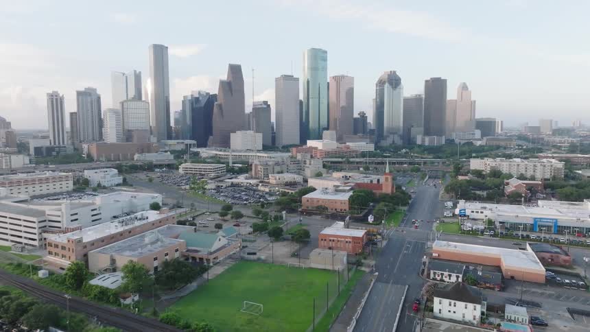 The video shows a highangle shot of a city with a skyline of tall buildings in the background in the foreground there are smaller buildings a parking lot and a grassy field a few cars can be seen driving on the roads