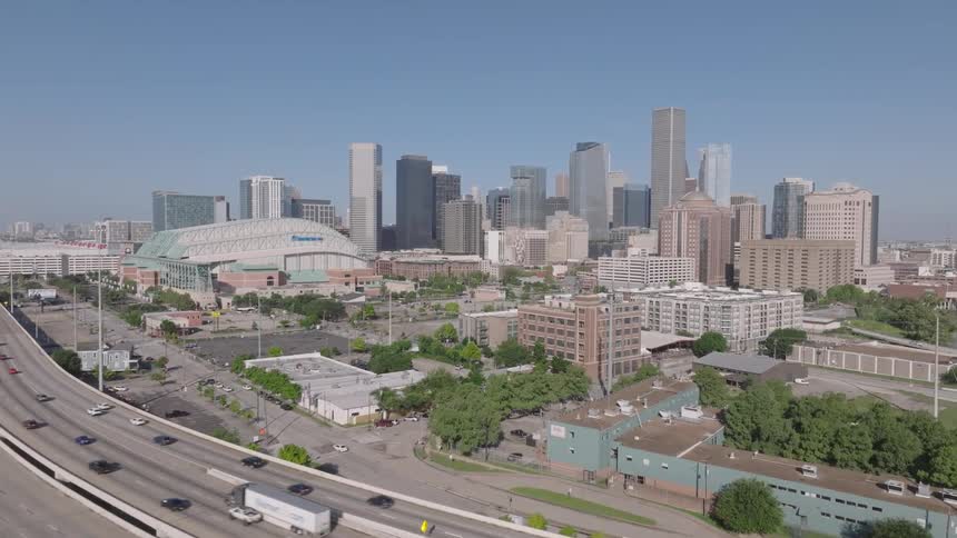 An aerial shot shows a busy freeway with cars driving on it leading towards a large city skyline on a clear day the city features many modern skyscrapers and a large stadium with a domed roof is prominent in the foreground