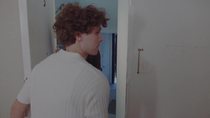 A shot begins with a view of a closed white door and then pans to the side as it opens a man with curly hair steps aside to reveal a woman standing in the doorway and the two briefly look at each other before the man turns away