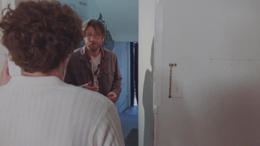 A medium shot shows a man with short hair and glasses walking into a room and talking to a man and woman in the doorway the man who just walked in gestures with his hands as he speaks while the other two listen