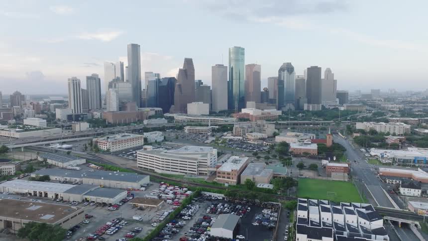 The video shows an aerial view of a city with a dense collection of skyscrapers in the background in the foreground there are smaller industrial and residential buildings along with a parking lot filled with cars and a small park