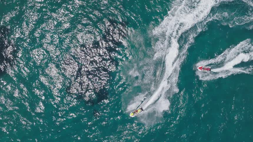Two jet skis leave white trails as they speed across the blue sea from above