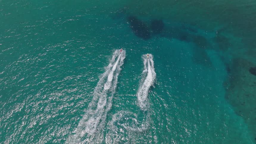 An aerial shot captures two people riding jet skis sidebyside in a turquoise ocean the jet skis are traveling away from the camera leaving white wakes in the clear water behind them