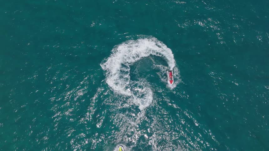 An aerial view shows a person on a red jet ski turning in a tight circle on the bright blue ocean the jet ski leaves a large white trail of foam and spray behind it and another jet ski is visible entering the frame