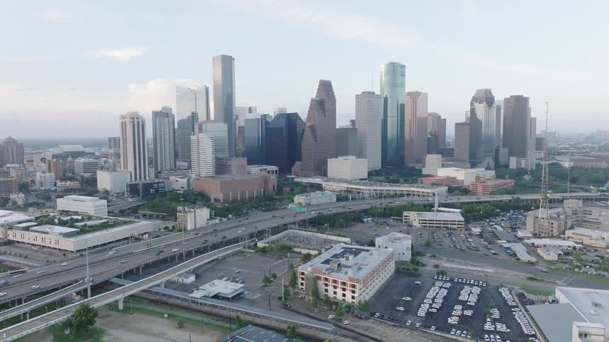 The video shows an aerial shot of a city with a skyline of tall buildings in the foreground there is a highway with cars driving on it and a large parking lot to the side the sky is hazy and overcast