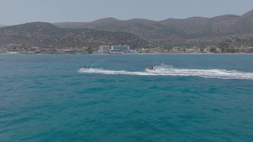 Two individuals on jet skis speed across a large body of blue water leaving a white wake behind them with a coastline of buildings and hills visible in the background