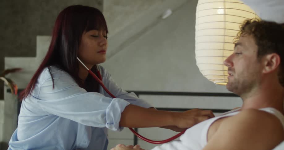 A woman uses a stethoscope to check the heartbeat of a young man lying on a bed wearing a singlet at midday