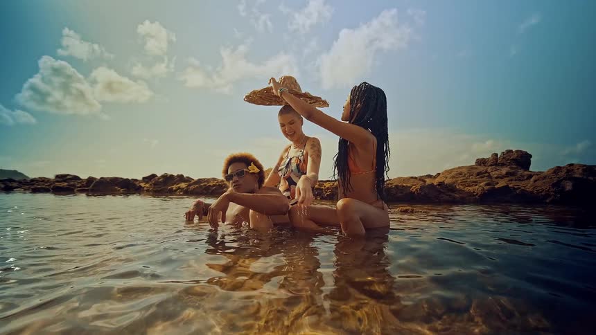 Three people bathing at the beach in a shallow water area two women and a man with one woman putting a beach hat on the other
