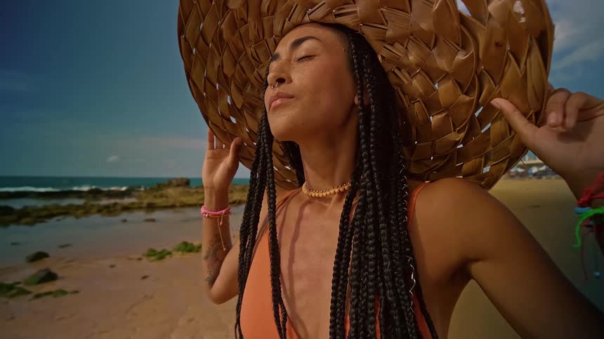 Close up portrait of a young woman holding her sunhat on her head while standing with her eyes closed outdoors at the beach during daytime