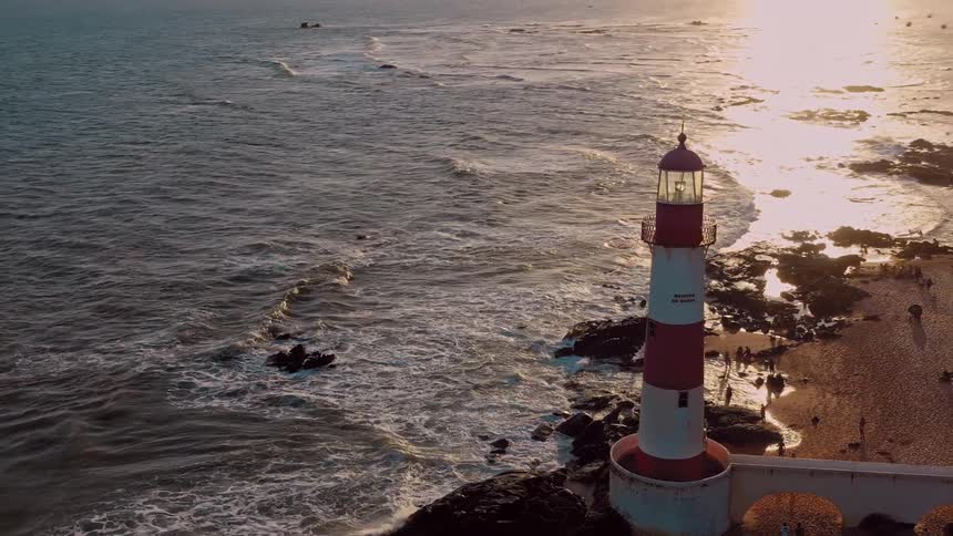 An aerial view shows a red and white striped lighthouse on a rocky coastline the sun is setting over the ocean casting a golden light on the waves and the beach