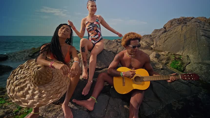 Three people sitting on a large rock at the beach with two women dancing and a man singing and playing the guitar