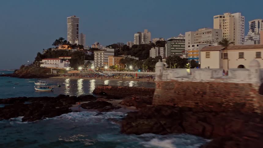 Historic and modern buildings blend by the seaside with boats and waves crashing against the rocks