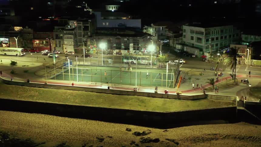 The video shows an elevated view of a city at night theres a welllit soccer field in the foreground with people playing and a river or canal flowing next to it in the background streetlights illuminate various buildings and cars on the road