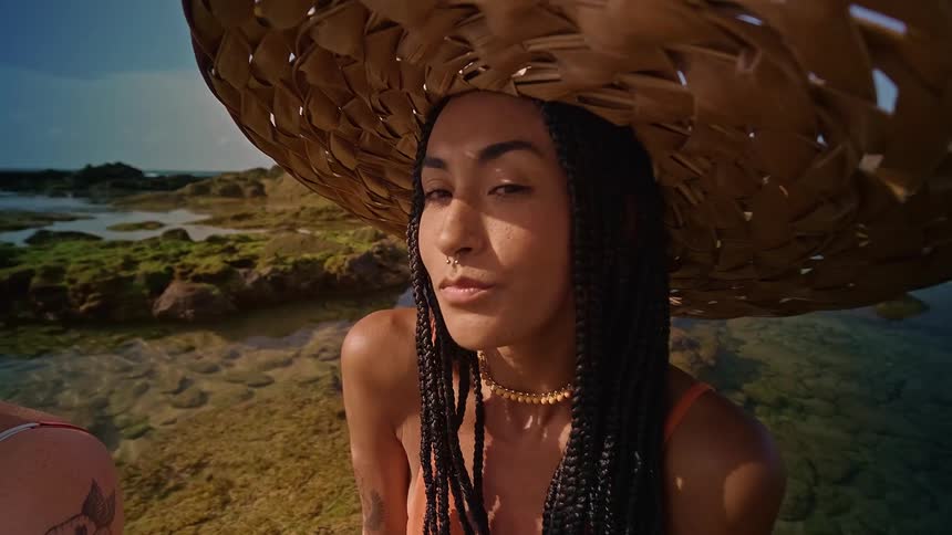A person with long braids a nose ring and a wide straw hat is sitting on a rocky beach near the ocean the camera zooms in on them as they turn their head from a profile to a headon view looking directly at the camera