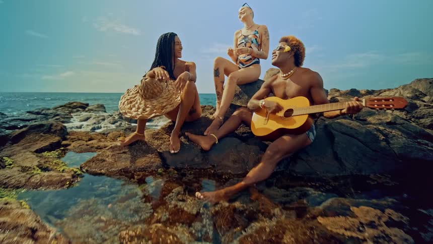 A man playing guitar strings while wearing sunglasses with two women sitting behind him on a rocky coast