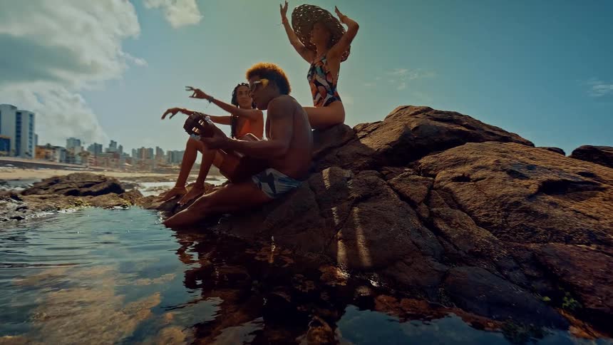 Three people in swimsuits sitting on seaside rocks one of them playing a guitar