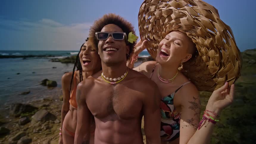 A carefree trio laugh together on the shore with one wearing a giant woven hat and another adorned with a flower and sunglasses
