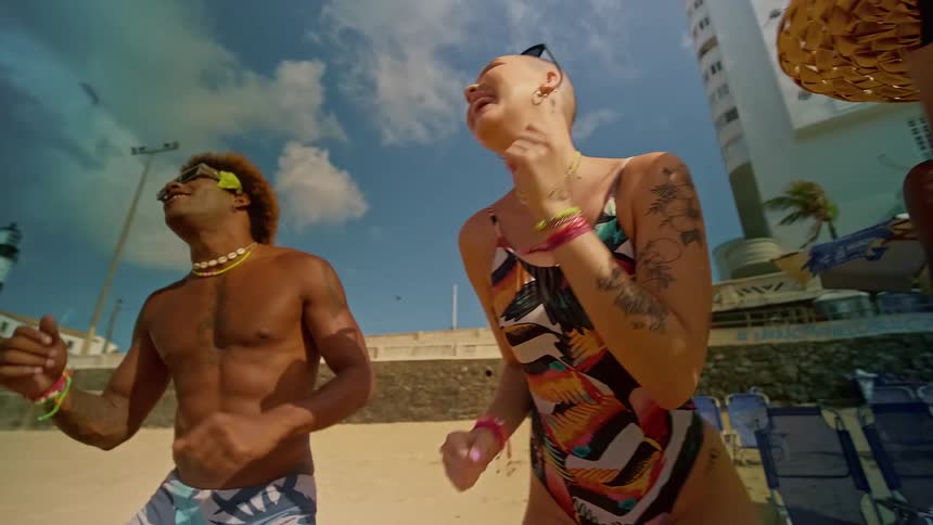 The video shows a group of three young people dancing on a sandy beach a lighthouse and a large white building can be seen in the background the person in the center is a person with a shaved head and tattoos wearing a colorful onepiece swimsuit