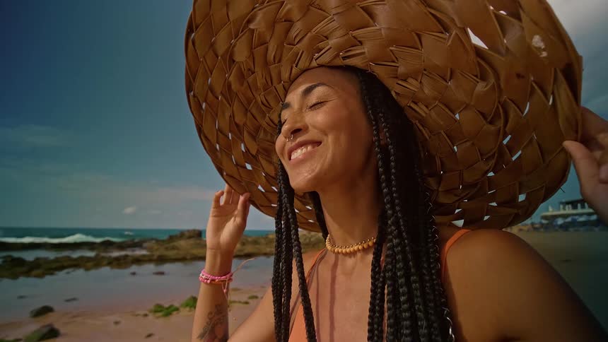 Close up portrait of a young woman holding her sunhat on her head while standing outdoors at the beach during daytime