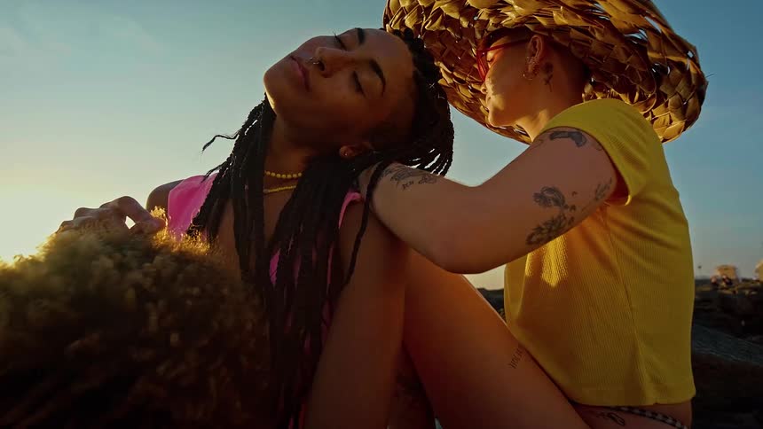A person with a large straw hat sits with their arm around a friend with long braids and a nose ring they are sitting close together with their eyes closed and heads tilted back enjoying the sunshine on a rocky beach
