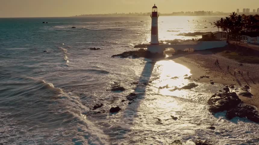 Waves crash against a rocky shoreline as the setting sun casts a long shadow of a coastal lighthouse across the shimmering ocean and sandy beach