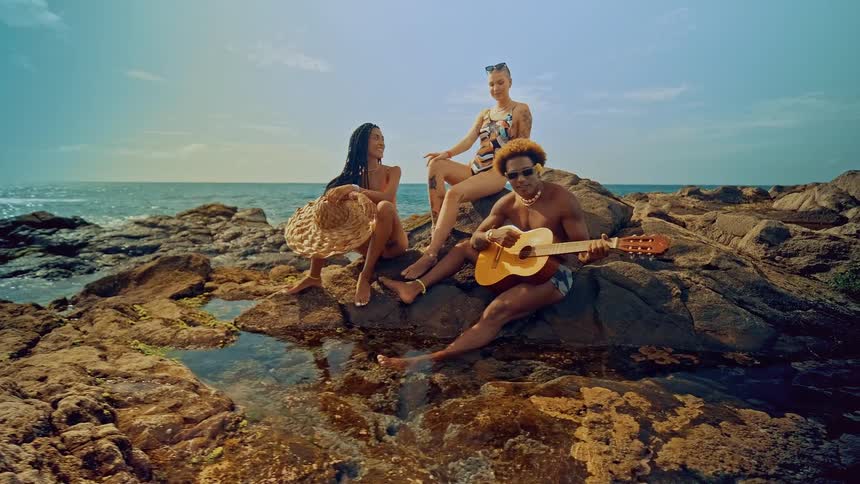 A group of three friends is sitting on the rocky shore by the ocean one person is playing a guitar while the others relax and enjoy the sunny day
