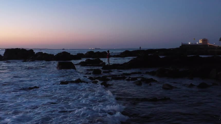 A lone person fishes on a rocky shoreline as waves break on the shore under a fading sunset with a small building and distant ships in the background