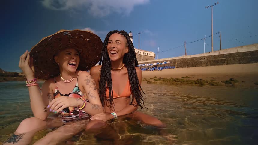 Two women in the shallow beach water one wearing a sun hat both laughing under the sun