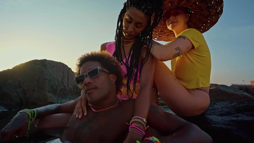 A man sitting in a pool of water wearing sunglasses with two womenone black and one whitesitting behind him on a rock at sunset
