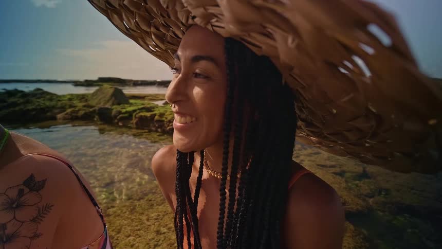 Two young women sitting on a rocky coastline at midday one with a tattooed arm and the other with braided hair wearing a large sun hat both smiling