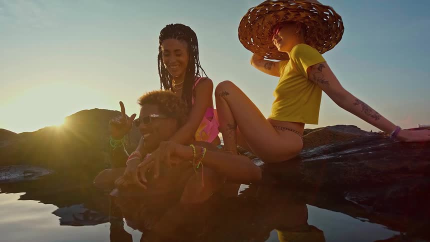 Three young adults two women and one man are relaxing by a natural body of water at sunset they are sitting on rocks that are partially submerged in the water with sunlight gleaming off the surface