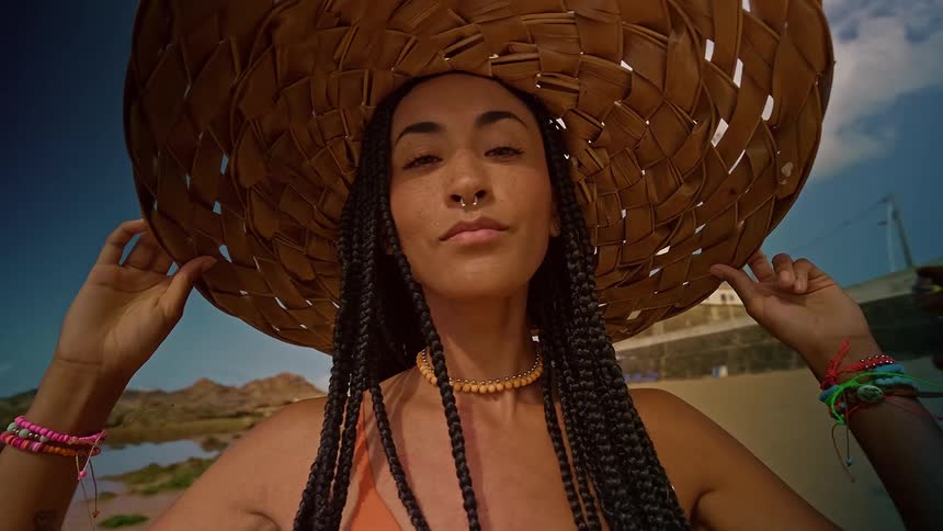 Close up portrait of a young woman holding her sunhat on her head while standing outdoors at the beach during daytime