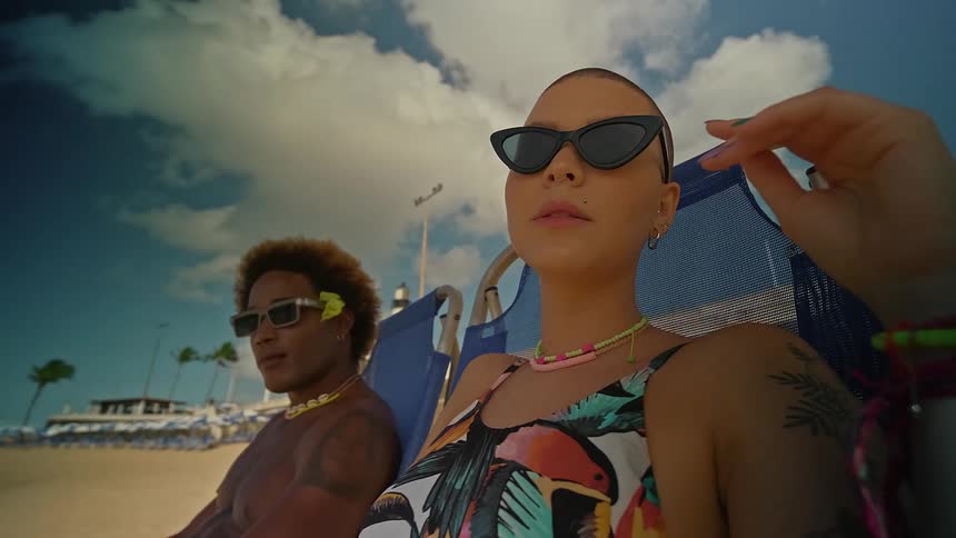 The video shows a man and a woman relaxing on the beach in what appears to be lounge chairs the man has brown curly hair and is wearing yellow sunglasses while the woman has a shaved head and is wearing black sunglasses they are both wearing colorful c